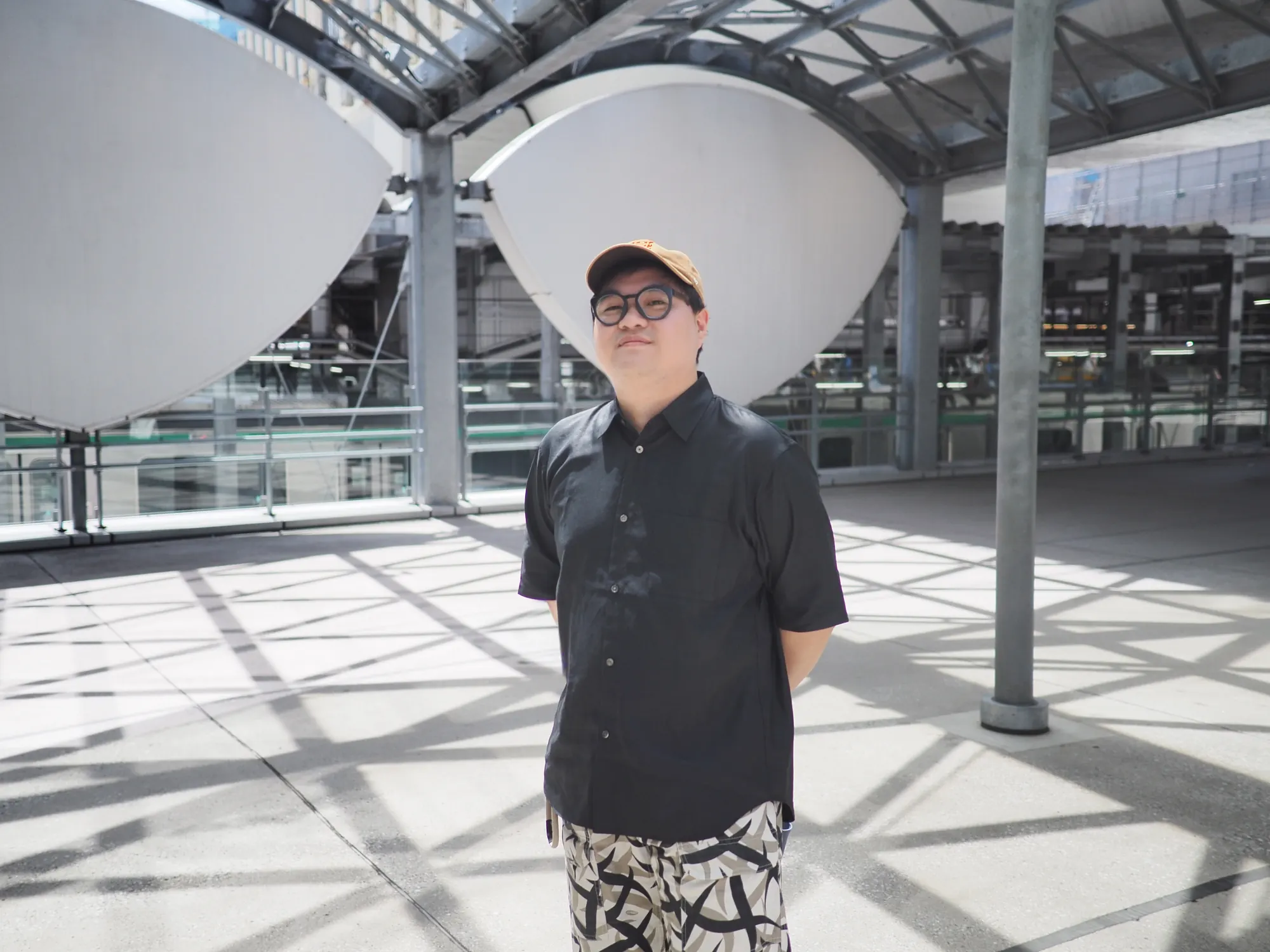 Munus Shih wearing glasses and a tan cap in a sunlit transit station